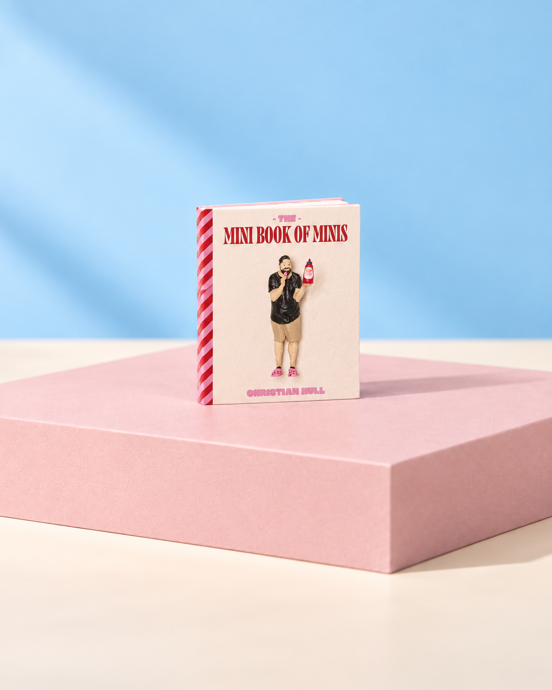 Mini Book of Minis with its Christian Hull cover, standing on a pink plinth against a blue background